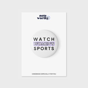 Könnte beinhalten: Weißer Knopf mit dem Text "WATCH WOMEN'S SPORTS" in Schwarz mit einem lila Streifen hinter dem Wort "WOMEN'S".