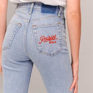 Op de afbeelding: Lichtblauwe denim jeans met een achterzak met de woorden "Raisin' Hell" geborduurd in rood.