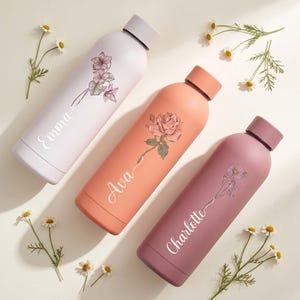 May include: Three pastel-coloured insulated water bottles. Each bottle has a floral design and a name in cursive script: Emma, Ava, and Charlotte. The colours are lavender, peach, and mauve. Daisies are scattered around.