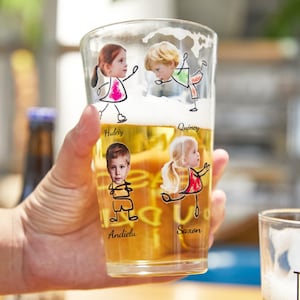 Puede incluir: Un vaso de pinta transparente lleno de cerveza, con ilustraciones de dibujos animados de niños con sus nombres impresos debajo. El vaso lo sostiene una persona. Las ilustraciones incluyen una niña con un vestido rosa, un niño con una camisa verde, un niño con una camisa naranja y una niña con un vestido rojo.