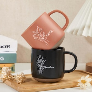 May include: Two ceramic mugs, one coral and one black, stacked on a wooden tray. The coral mug has "MAMA" and a floral design. The black mug has "Grandma" and a floral design. Both mugs have a brown base and a curved handle.