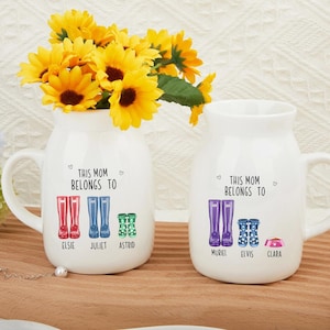 May include: Two white ceramic mugs with handles. Each mug has a design of colorful rain boots with names printed below. The text on the mugs reads "This Mom Belongs To".