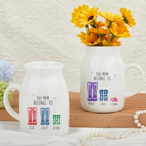 May include: Two white ceramic vases with handles, each featuring the text "THIS MOM BELONGS TO" and illustrations of boots. One vase has Elsie, Juliet, and Astrid's names, the other has Muriel, Elvis, and Clara's names. Yellow flowers are in one vase.