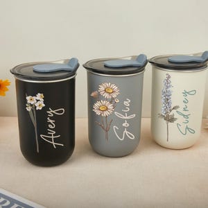 Könnte beinhalten: Drei personalisierte Reisebecher mit Blumendesigns. Die Becher sind schwarz, grau und cremefarben, jeder mit einer anderen Blume und einem Namen in Schreibschrift: Avery, Sofia und Sidney. Jeder Becher hat einen schwarzen Deckel mit einem blauen Schieber.