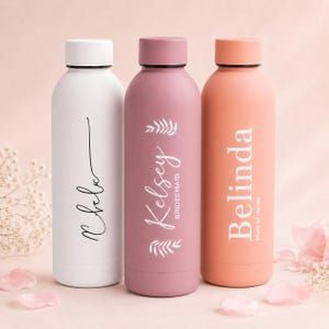 Op de afbeelding: Drie cilindrische metalen waterflessen met schroefdoppen. De witte fles heeft zwarte cursieve tekst, de mauve fles toont witte tekst met Kelsey Bridesmaid en bladaccenten, en de perzikkleurige fles toont witte tekst met Belinda Maid of Honor.