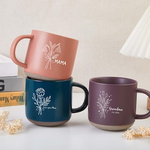 May include: Three ceramic mugs in shades of pink, teal, and purple. Each mug features a floral design and text: "Mama", "Love you Mom", and "Grandma Est. 2026". The mugs have a speckled texture and a contrasting base.