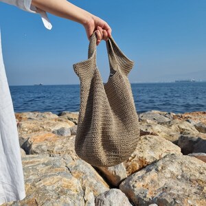 May include: A beige crocheted tote bag with a wide opening and thick straps. The bag is held by a hand against a backdrop of rocks and blue water.
