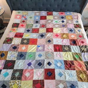 May include: A patchwork quilt with a variety of colorful fabric squares, each with a contrasting diamond-shaped accent. The quilt is spread across a bed with a gray upholstered headboard, showcasing a vintage, handmade aesthetic.