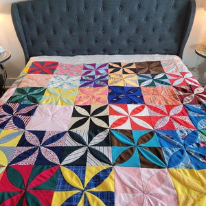 May include: A colorful patchwork quilt with a pinwheel design. The quilt features squares of various fabrics in shades of red, blue, yellow, pink, and green. The quilt is placed on a bed with a gray upholstered headboard.