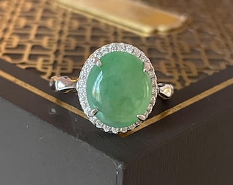 May include: A silver ring featuring a large, oval-shaped, light green gemstone, surrounded by a halo of small, clear stones. The ring is set on a dark surface, showcasing the bright gemstone.