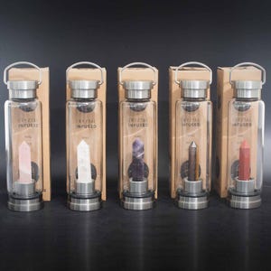 May include: Five glass water bottles with crystal infused stoppers. The bottles are clear glass with stainless steel lids and bases. The crystals are pink, white, purple, brown, and red. The bottles are labelled "Crystal Infused".
