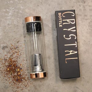 May include: A glass water bottle with a rose gold lid and a built-in infuser. The bottle is sitting on a grey surface with loose tea leaves scattered around it. The bottle is labelled "CRYSTAL BOTTLE".