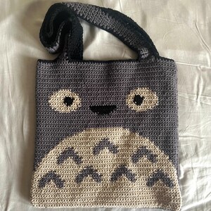 May include: A crocheted tote bag with a gray and white Totoro design. The bag has black straps and a smiling face.