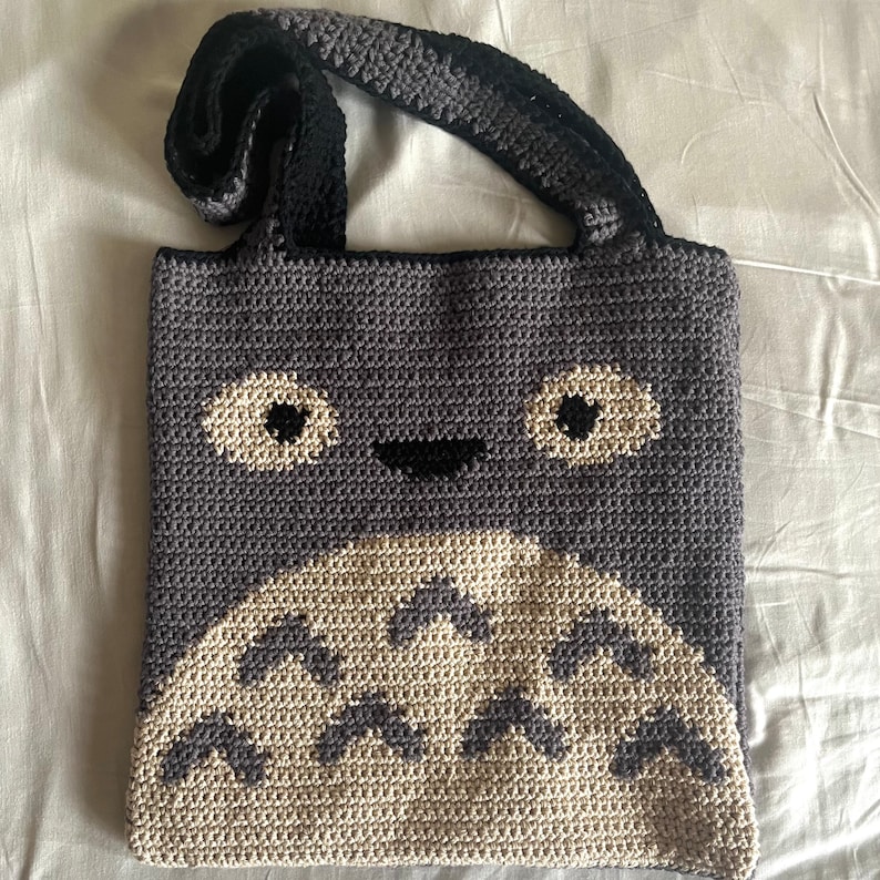 May include: A crocheted tote bag with a gray and white Totoro design. The bag has black straps and a smiling face.