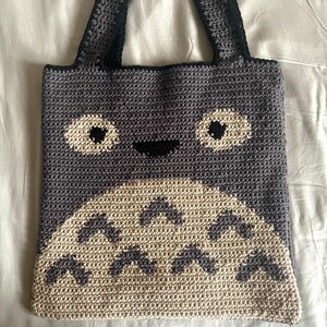 May include: A gray crocheted tote bag with a white and black Totoro design. The bag has two black straps.