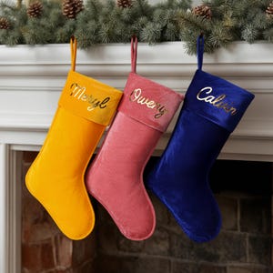 Peut inclure: Trois bas de Noël en velours jaune, rose et bleu sont suspendus à une cheminée. Chaque bas a un revers doré avec un nom brodé en lettres dorées : Meryl, Owen et Calvin. Les bas sont suspendus par des boucles en tissu assorties.