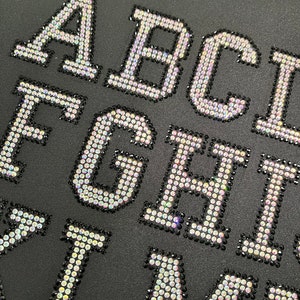 May include: Close-up of a black fabric with the letters A, B, C, D, E, F, G, H, I, J, K, L, M, N, O, P, Q, R, S, T, U, V, W, X, Y, and Z spelled out in iridescent rhinestones.
