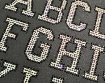 May include: Close-up of a black fabric with the letters A, B, C, D, E, F, G, H, I, J, K, L, M, N, O, P, Q, R, S, T, U, V, W, X, Y, and Z spelled out in iridescent rhinestones.