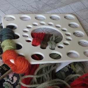 May include: A white plastic thread organiser with multiple holes, holding various colours of embroidery floss. Colours include red, green, and blue. The image also shows spools of thread and a cross-stitch pattern.