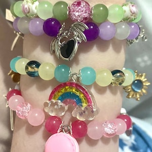 Pode incluir: Quatro pulseiras de contas coloridas com pingentes. A pulseira superior tem contas verde claro, roxo e branco com um pingente de dragão prateado. A segunda pulseira tem contas verde claro, azul e amarelo com um pingente de arco-íris prateado. A terceira pulseira tem contas azul claro, amarelo e verde com um pingente de sol dourado. A pulseira inferior tem contas rosa, vermelho e branco com um pingente de macaron rosa.