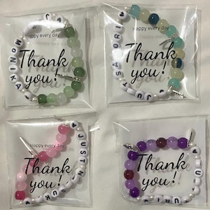 Puede incluir: Cuatro bolsas de plástico transparentes que contienen cada una un brazalete de cuentas con cuentas de letras blancas que deletrean un nombre y la frase "¡Gracias!" Los brazaletes están hechos con cuentas de diferentes colores: verde, rosa, morado y azul. Cada bolsa tiene una tarjeta pequeña con la frase "Feliz cada día" impresa en ella.