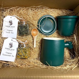 Puede incluir: Una caja de regalo llena de artículos relacionados con el té. Incluye una taza verde oscuro, un platillo y una maceta pequeña, junto con frascos de té de hojas sueltas y una cuchara de madera. Dos etiquetas dicen "Loose Leaf Tea Mug" con "Organic Nettle Tea" y "Organic Chamomile Tea".