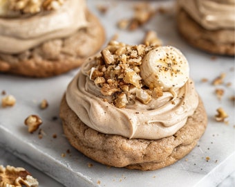 May include: Close-up of a brown sugar cookie topped with a thick layer of brown frosting, chopped walnuts, and a slice of banana. The cookie is sitting on a white marble surface.