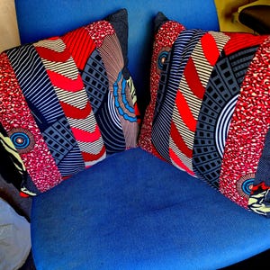 May include: Two patterned cushions with a patchwork design. The cushions feature red, black, grey, and white geometric and abstract patterns. They are placed on a blue chair, highlighting their colourful design.