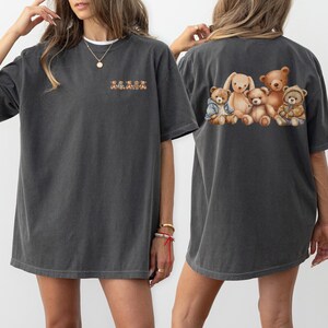 Puede incluir: Una camiseta gris oscuro de gran tamaño con un gráfico de cinco osos de peluche en la espalda. Los osos llevan diferentes atuendos, incluyendo una chaqueta vaquera y una pajarita.