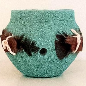 May include: A turquoise textured pot with a granular surface finish. The vessel features two dark brown, feather-like decorative accents on the sides, each secured with a thin, light-colored cord. A small, circular hole is visible in the center.