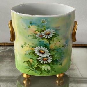 May include: A cylindrical ceramic vase with a hand-painted floral design featuring white daisies with yellow centers and green leaves. The vase has gold-colored handles and feet, and a pastel green, yellow, and blue background.