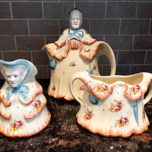May include: A vintage ceramic tea set featuring a teapot, creamer, and sugar bowl. The set is decorated with floral patterns and a woman's face on each piece. The colors include cream, blue, pink, and orange.