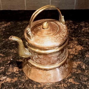 May include: A vintage copper teapot with an ornate engraved design. The teapot has a brass handle and spout, and a lid with a finial.