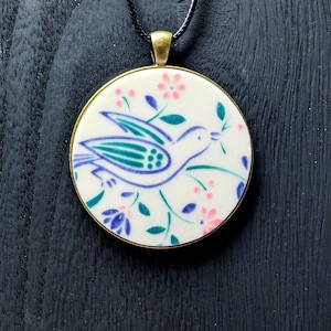 May include: A white ceramic pendant necklace with a blue and green bird design and floral accents. The pendant is round and has a gold-colored metal frame. The necklace has a black cord.