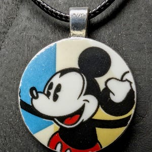May include: A round pendant featuring Mickey Mouse holding an apple. The pendant has a silver bail and hangs on a black cord. The background is divided into blue, white, and yellow sections.