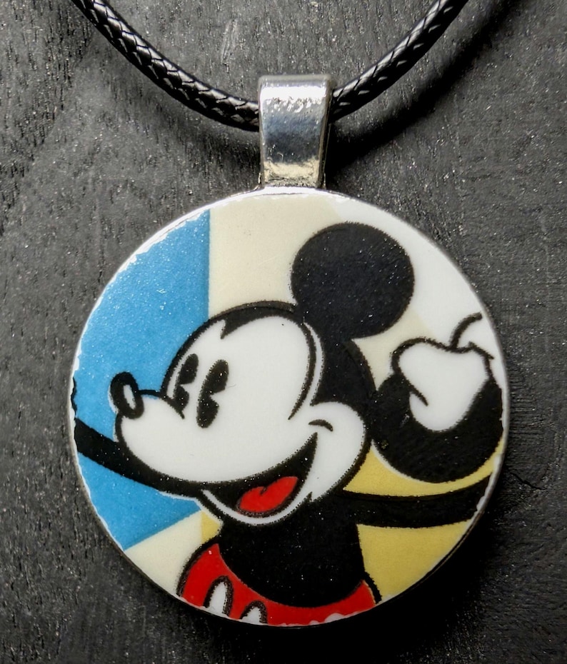 May include: A round pendant featuring Mickey Mouse holding an apple. The pendant has a silver bail and hangs on a black cord. The background is divided into blue, white, and yellow sections.