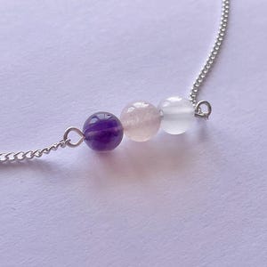 May include: A delicate necklace featuring three gemstone beads: a deep purple amethyst, a soft pink rose quartz, and a translucent white moonstone. The beads are strung on a fine silver chain, creating a minimalist and elegant design.