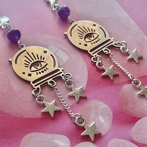 Può includere: Orecchini color argento con un design a sfera di cristallo, un occhio e un dettaglio a raggiera. Ogni orecchino ha una perla viola e stelle pendenti. Gli orecchini sono esposti su una superficie rosa con pietre rosa chiaro.