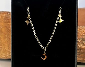 Könnte beinhalten: Eine goldfarbene Halskette mit Kette und drei Anhängern: ein Halbmond und zwei Sternschnuppen. Die Halskette wird in einer schwarzen Schachtel mit Holzgrund präsentiert.