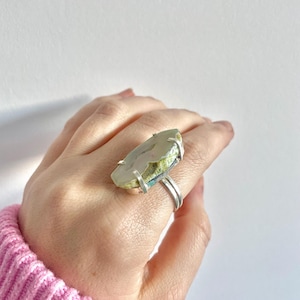 May include: A silver ring featuring a large, green, white and blue agate stone set in the centre. The ring is worn on a person's finger.