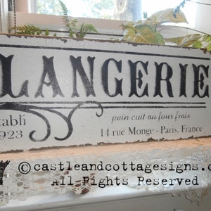 May include: A distressed white wooden sign with the word "BOULANGERIE" in large black letters. Below, it reads "&egrave;tabli 1923" and "pain cuit au four frais, 14 rue Monge - Paris, France". The sign has a vintage, rustic aesthetic.