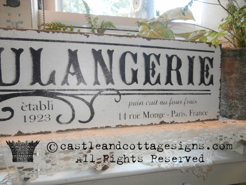 May include: A distressed white wooden sign with the word "BOULANGERIE" in large black letters. Below, it reads "&egrave;tabli 1923" and "pain cuit au four frais, 14 rue Monge - Paris, France". The sign has a vintage, rustic aesthetic.