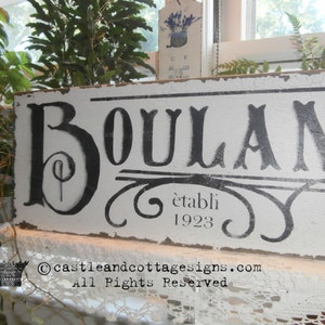 May include: A distressed white wooden sign with the word "BOULANGERIE" in large black letters. The sign also includes the text "&eacute;tabli 1923" and decorative flourishes. The sign is placed near plants and a window.