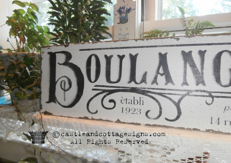 May include: A distressed white wooden sign with the word "BOULANGERIE" in large black letters. The sign also includes the text "&eacute;tabli 1923" and decorative flourishes. The sign is placed near plants and a window.
