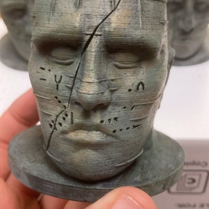 以下が含まれることがあります： グリッドパターンと側面にひびが入った、グレーとグリーンの3Dプリントされた人間の頭部の胸像。胸像は丸い台座の上に置かれています。