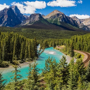May include: A scenic view of a turquoise river winding through a lush green forest, with snow-capped mountains in the background. A train track runs along the edge of the river.