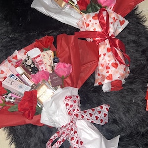 May include: Bouquets of beauty products, including perfumes, lip glosses, and lotions, are arranged with red and pink roses. The bouquets are wrapped in white tissue paper and red paper, tied with ribbons featuring heart patterns.