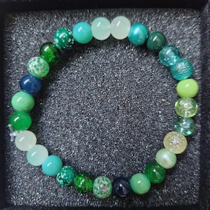 Puede incluir: Un brazalete de cuentas verdes y azules. Las cuentas son de varios tonos y texturas, incluyendo moteadas y translúcidas. El brazalete se presenta sobre una superficie oscura. Las cuentas son de diferentes tamaños.