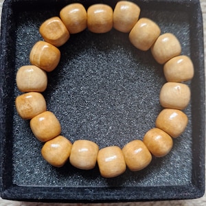 Può includere: Un braccialetto di perline di legno marrone chiaro. Il braccialetto è composto da perline lisce di forma quadrata infilate insieme. Le perline hanno una venatura naturale del legno e sono esposte in un vassoio quadrato nero.