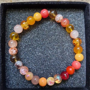 May include: A bracelet made of colourful, round gemstones in shades of orange, yellow, pink, and red. The beads are strung together. The bracelet is presented on a black, textured surface.